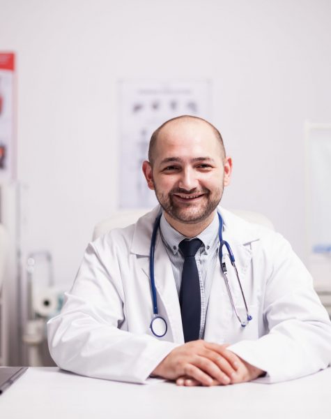 Portrait of confident medical practitioner in hospital office smiling at camera dressed with white coat and stethoscope around his neck.